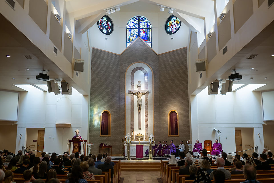 A church interior with a crucifix, stained glass windows, and people in pews. A speaker stands at a pulpit, and clergy in purple sit nearby.