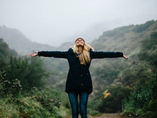 Woman in a dark coat stands with arms outstretched in a foggy, lush green mountain setting, expressing joy and freedom.