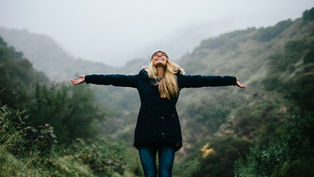 Woman in a dark coat stands with arms outstretched in a foggy, lush green mountain setting, expressing joy and freedom.