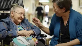 Elderly woman in wheelchair smiles at woman in blue cardigan touching her hand. They're in a hallway, conveying warmth and connection.