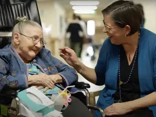 Elderly woman in wheelchair smiles at woman in blue cardigan touching her hand. They're in a hallway, conveying warmth and connection.