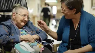 Elderly woman in wheelchair smiles at woman in blue cardigan touching her hand. They're in a hallway, conveying warmth and connection.