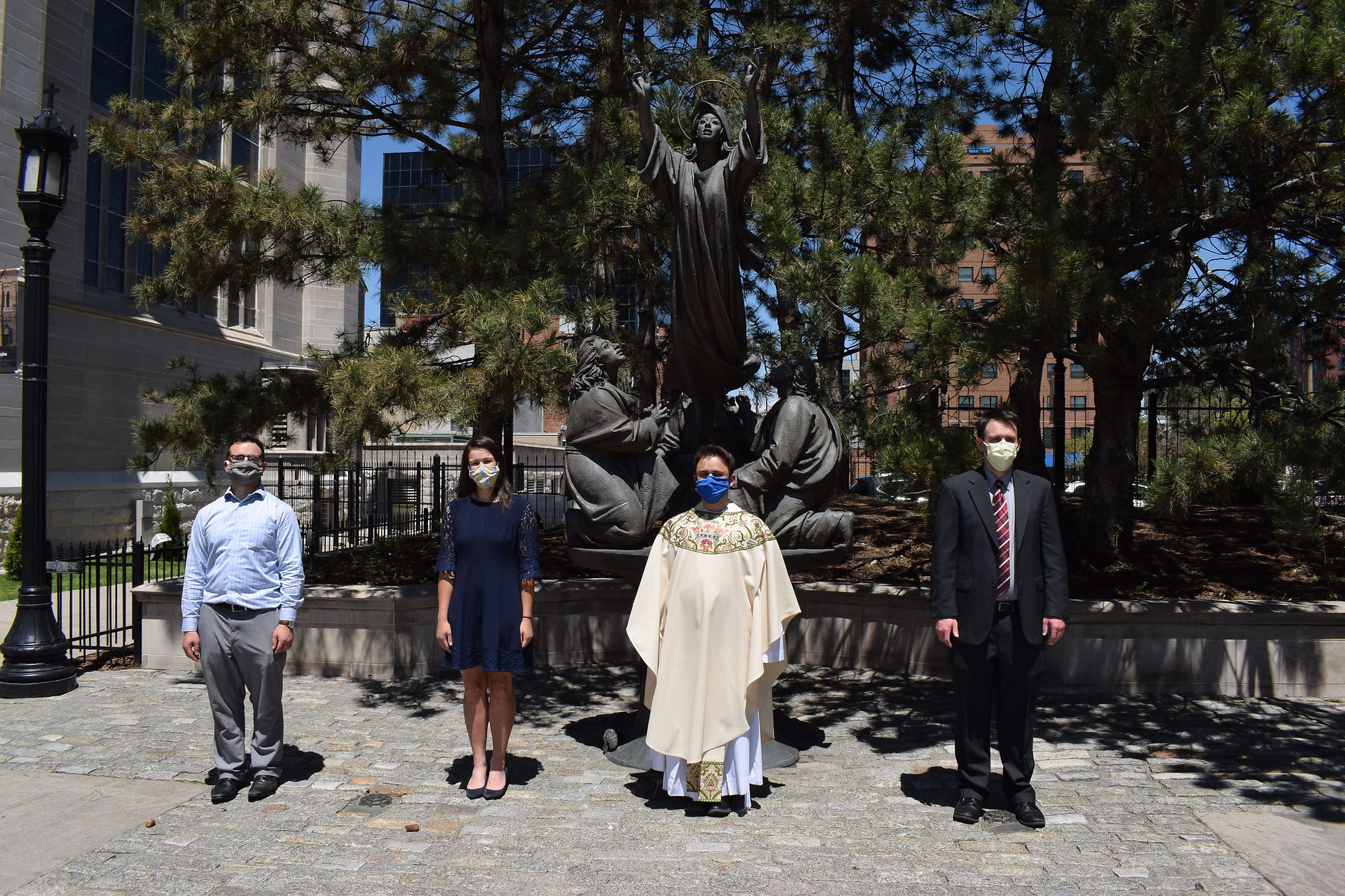 Four people wearing masks stand in front of a religious statue outdoors, on a sunny day by a building. Trees and cityscape in background.