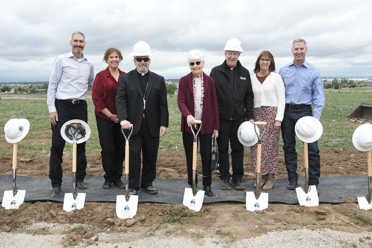 Seven people with shovels stand together at a groundbreaking event. They're outside on a cloudy day, wearing business casual attire and hard hats.