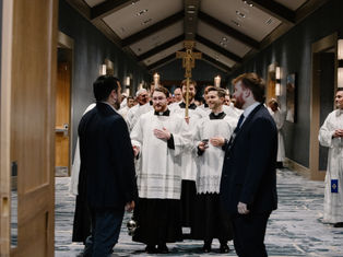 Catholic seminarians in white vestments walk in procession down a hallway, carrying candles and a crucifix, as attendees stand on either side greeting them.