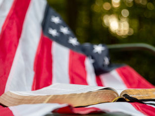 Open Bible resting on a draped American flag, with a soft outdoor bokeh background and a reflective, patriotic mood