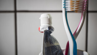 Toothpaste tube and two colorful toothbrushes in a cup against a tiled bathroom wall.