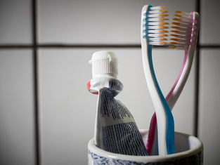 Toothpaste tube and two colorful toothbrushes in a cup against a tiled bathroom wall.