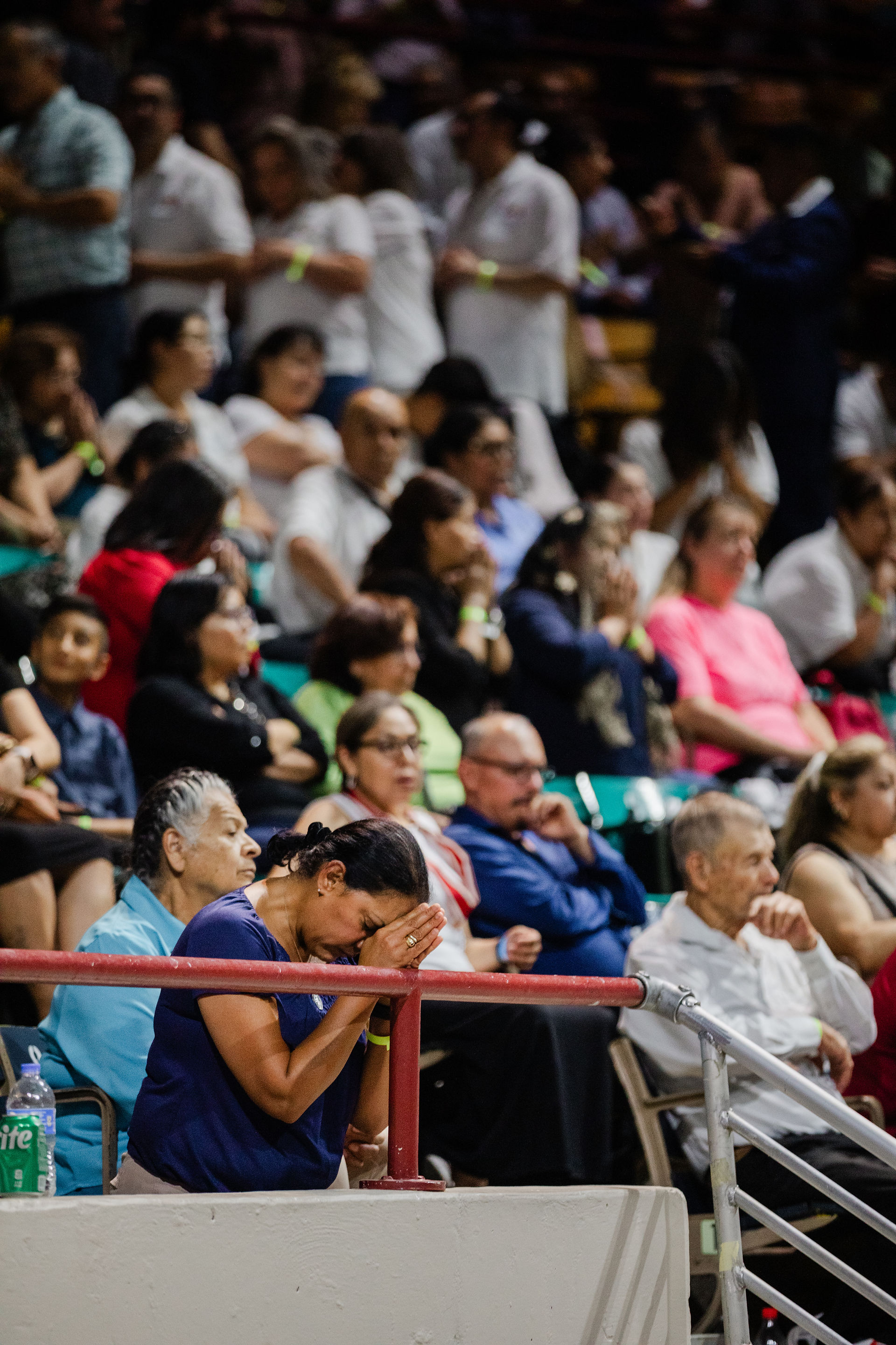 A large crowd seated in an indoor arena; people are talking or observing, with a red railing in front and a Sprite bottle visible on the left.