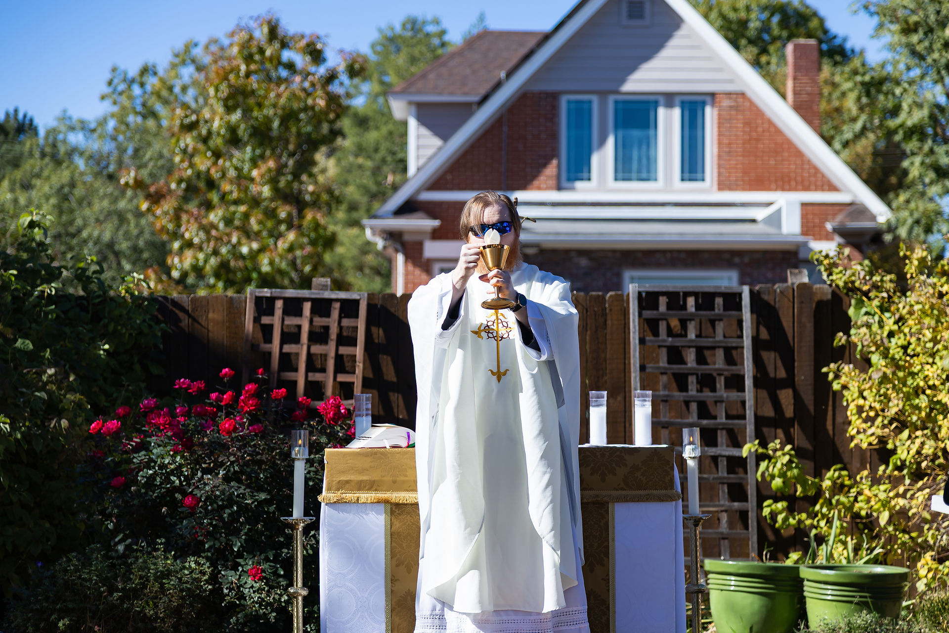 Priest in white robe holds a chalice outdoors, with red flowers and a house in the background. Bright, sunny day creates a serene mood.