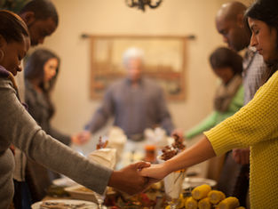 Six people hold hands in prayer around a dining table set with decorations. Warm, cozy atmosphere with focus on togetherness.