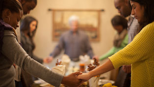 Six people hold hands in prayer around a dining table set with decorations. Warm, cozy atmosphere with focus on togetherness.