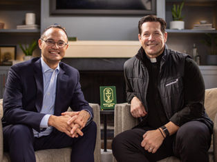 Two men smiling, sitting on a sofa in a cozy room. One in a suit, the other in a clerical collar and black vest. Green book on the table.