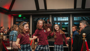 Three smiling girls in plaid skirts and maroon tops dance joyfully indoors. People seated in the background watch and applaud.