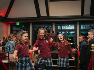 Three smiling girls in plaid skirts and maroon tops dance joyfully indoors. People seated in the background watch and applaud.