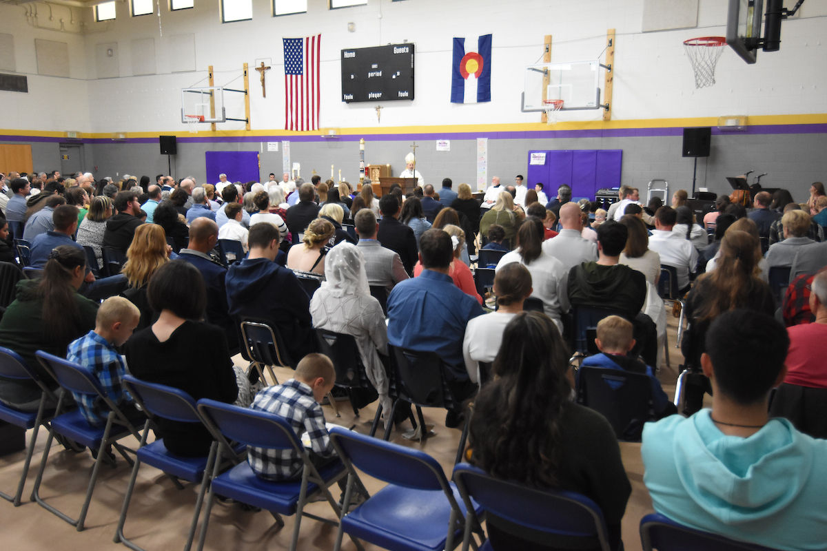 Audience seated in a gym watching a speaker at a podium. Flags and scoreboard in background. People attentive, various ages present.