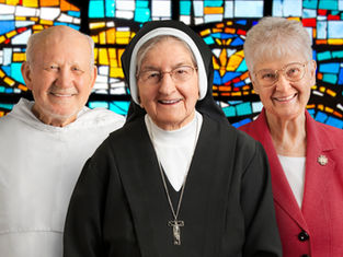 Three elderly people smiling, a priest, a nun, and a woman in a pink blazer. Colorful stained glass background enhances warmth.