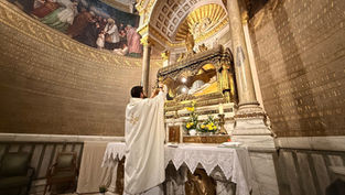 Priest in white robe holds up a chalice at ornate altar with candles, flowers, and gold details. Mural and inscriptions adorn the walls.