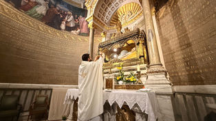 Priest in white robe holds up a chalice at ornate altar with candles, flowers, and gold details. Mural and inscriptions adorn the walls.