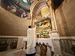 Priest in white robe holds up a chalice at ornate altar with candles, flowers, and gold details. Mural and inscriptions adorn the walls.