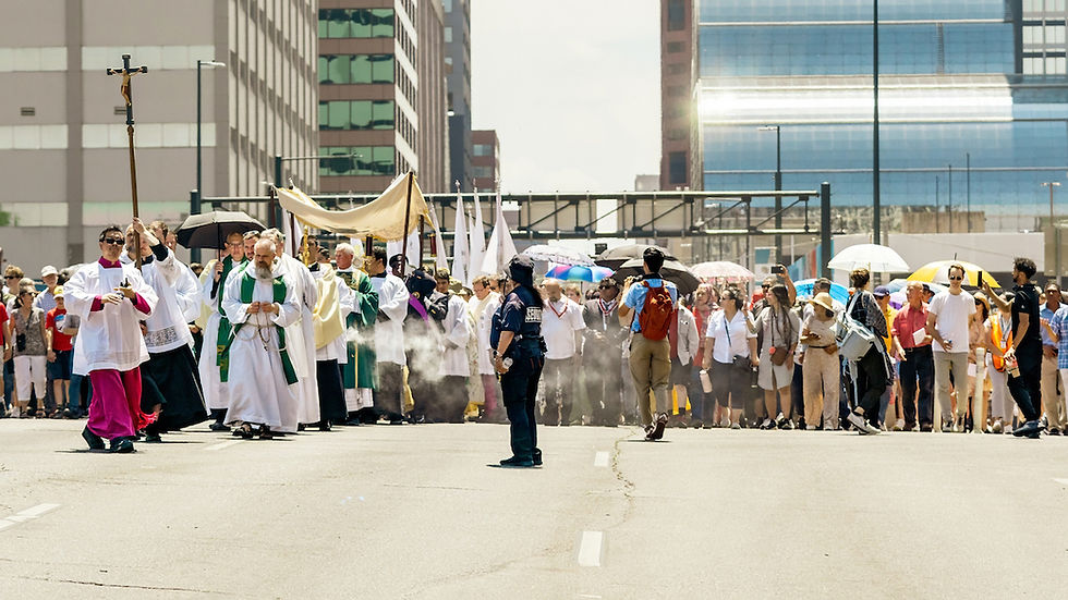 Priests in ceremonial robes lead a procession down a city street. A crowd follows, some with umbrellas. A security officer stands by.