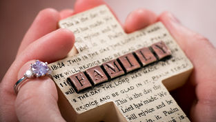 Hands holding a beige cross embossed with text and "FAMILY" in brown blocks. A ring with a sparkling stone is on one finger.