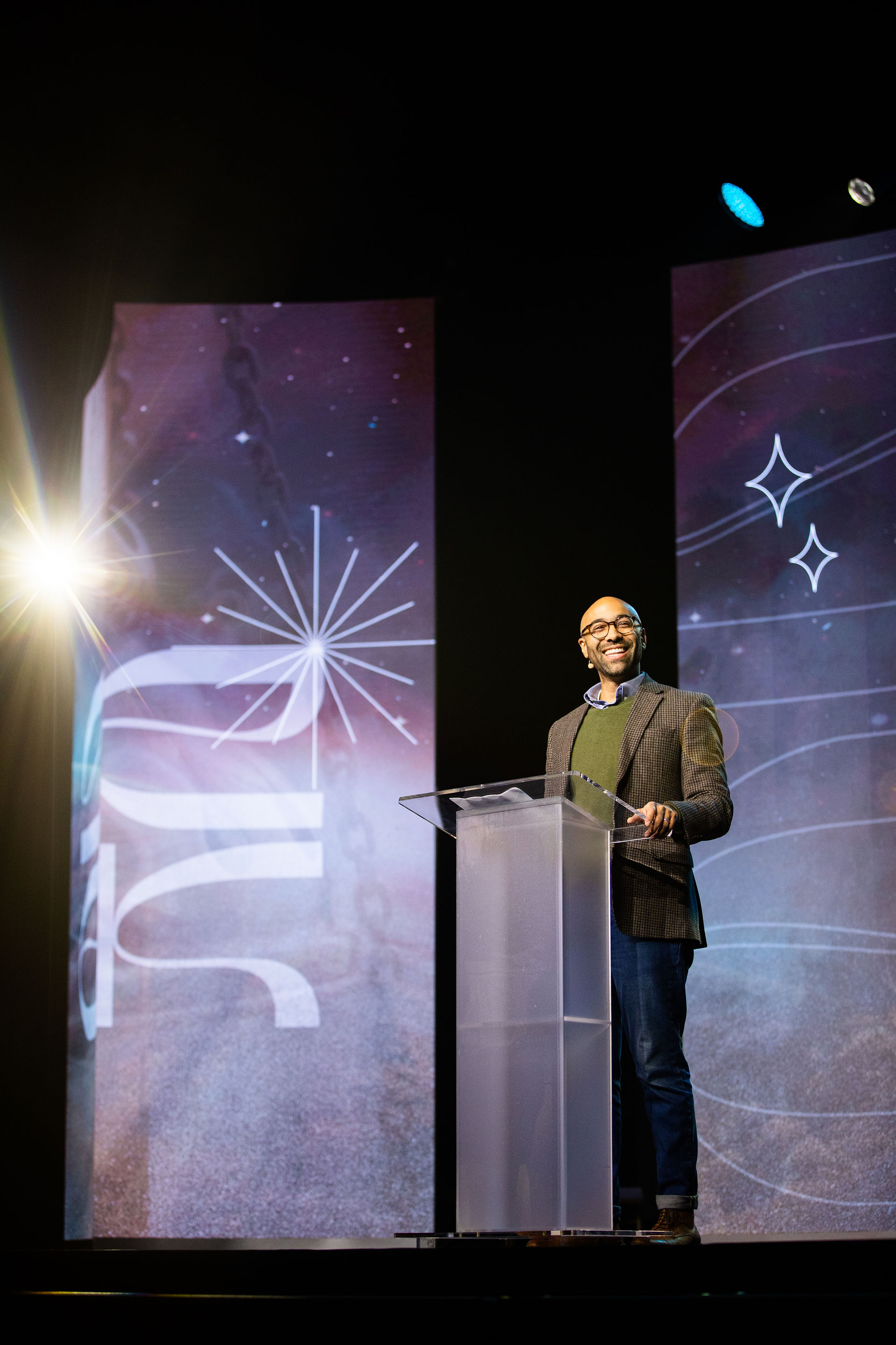 Man speaking at a podium on stage, smiling. Abstract star patterns in the background with a spotlight on the left. Dark setting.