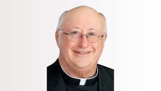 Smiling bald man in clergy attire with glasses, against a white background. He wears a black robe with a white clerical collar.