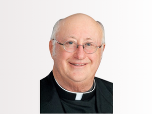 Smiling bald man in clergy attire with glasses, against a white background. He wears a black robe with a white clerical collar.