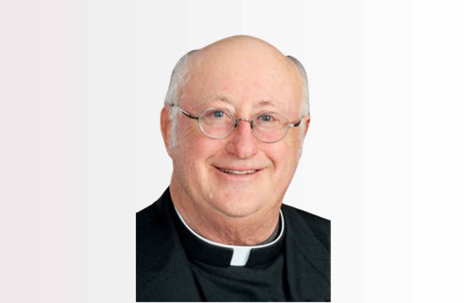 Smiling bald man in clergy attire with glasses, against a white background. He wears a black robe with a white clerical collar.