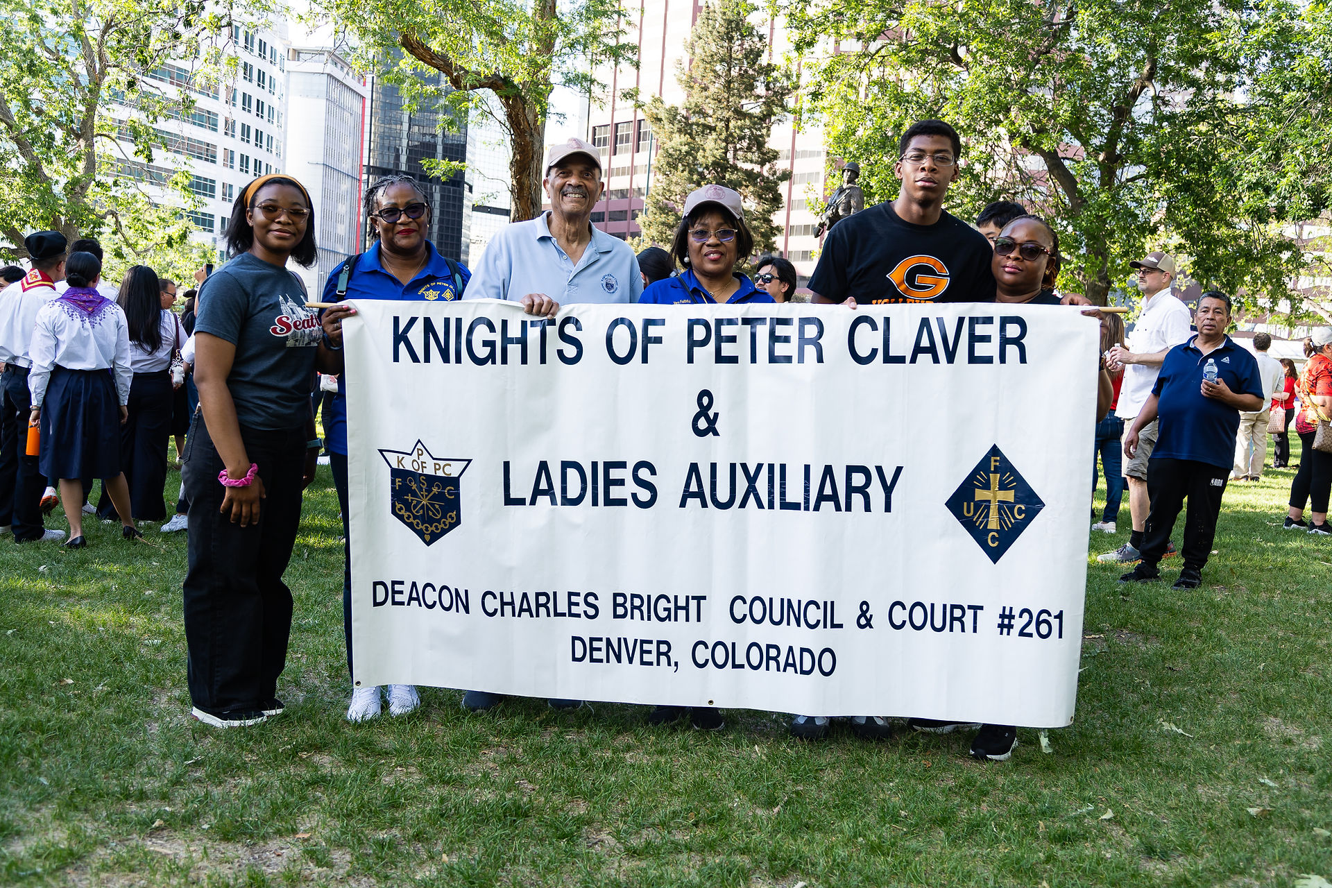 Group of people in a park hold a banner reading Knights of Peter Claver and Ladies Auxiliary, smiling during a community gathering.
