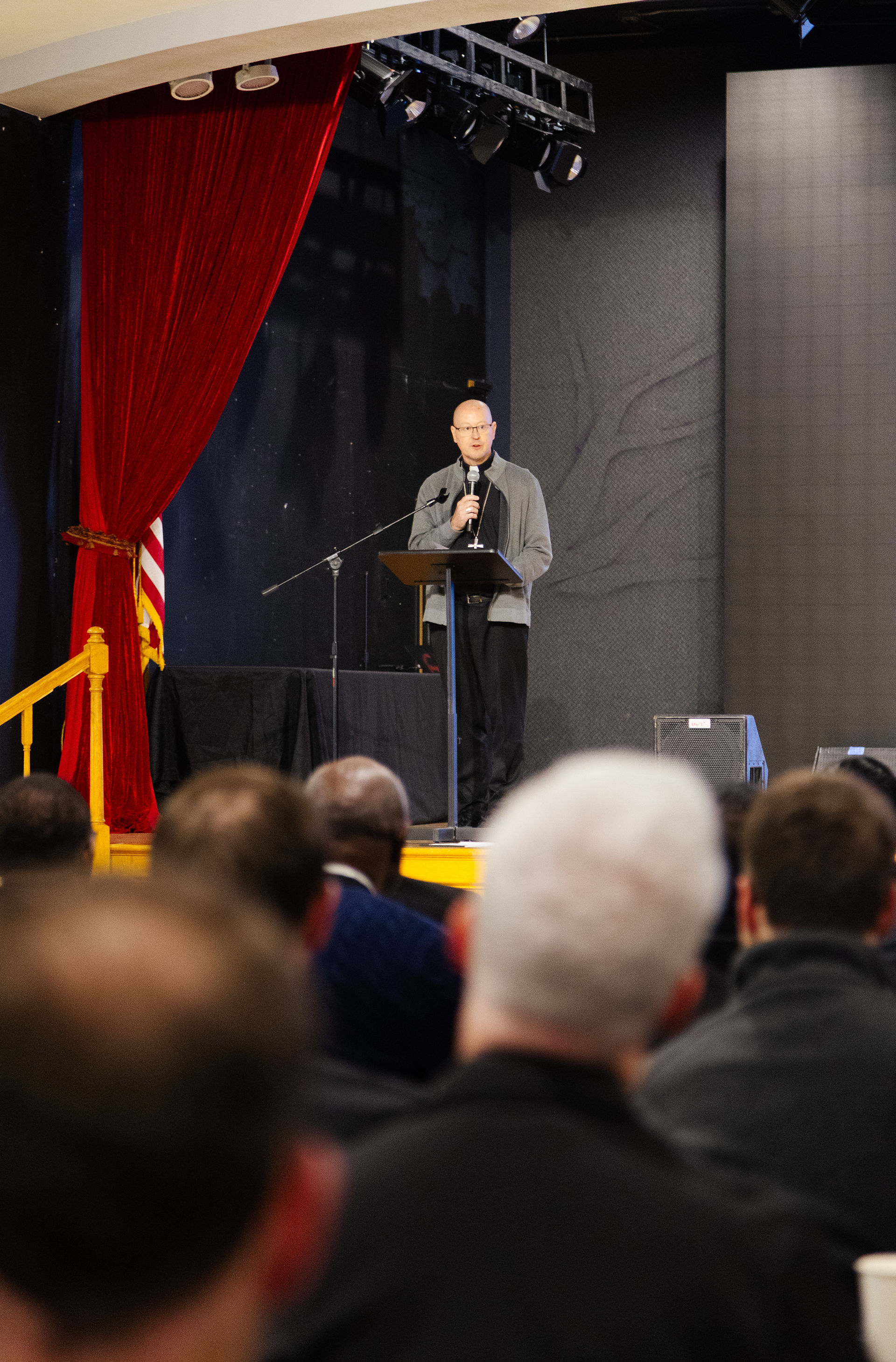A person speaks at a podium on stage with a red curtain, addressing an audience. The background is dark, creating a formal atmosphere.