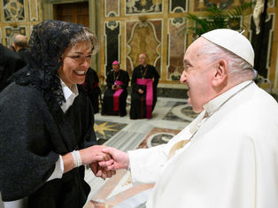 A smiling woman in a black lace veil shakes hands with a man in white in an ornate room. Two men in black robes and pink sashes sit in the background.
