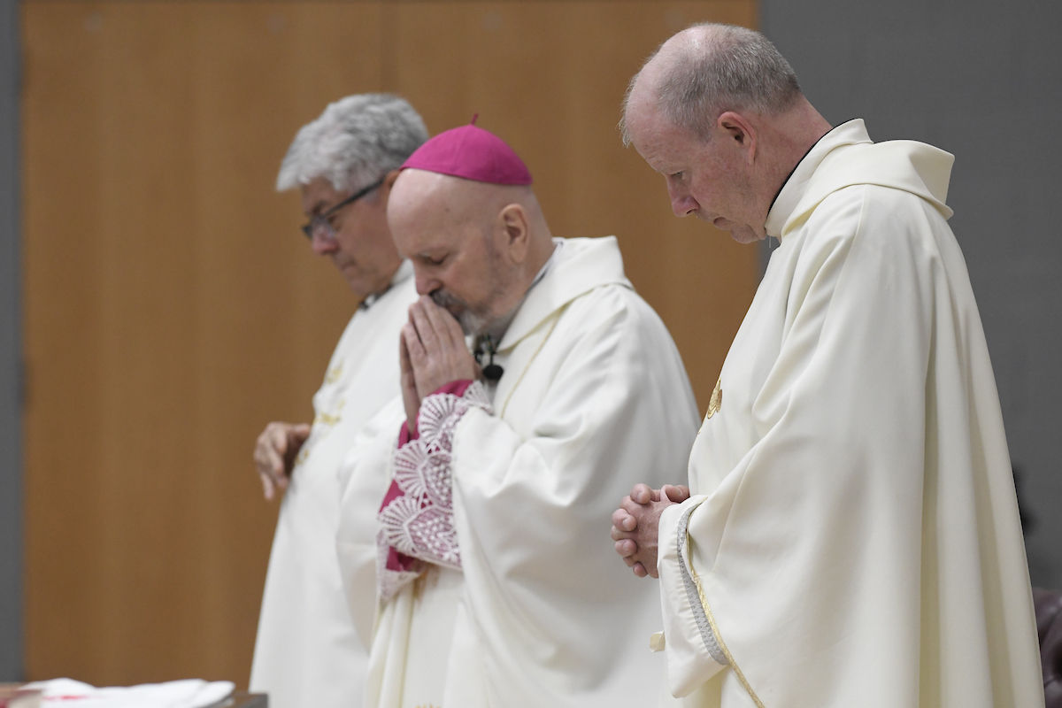 Three clergymen in white robes stand in prayer, eyes closed, hands clasped. Wooden wall background, solemn mood.
