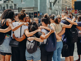 A group of people huddled in an embrace outdoors in an urban setting. They display unity and support. Buildings and signage in the background.