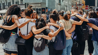 A group of people huddled in an embrace outdoors in an urban setting. They display unity and support. Buildings and signage in the background.