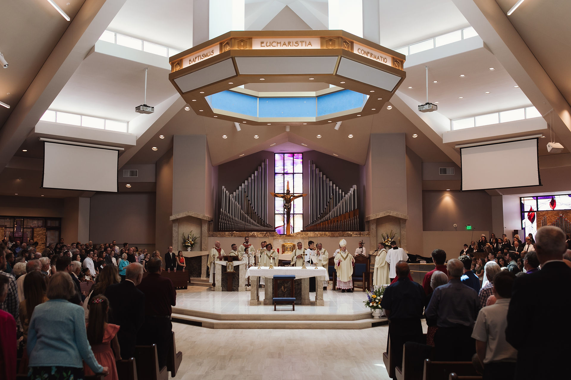 Church service with priests at the altar, surrounded by a congregation. Large organ, stained glass, and "Eucharistia" seen above.