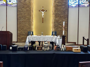 Church altar with a white cloth, crucifix on brick wall, and various urns and boxes on a table in front. Stained glass windows above.
