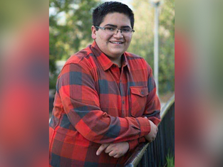 Smiling person in glasses and red plaid shirt leans on railing outdoors, with blurred greenery in the background, creating a relaxed mood.