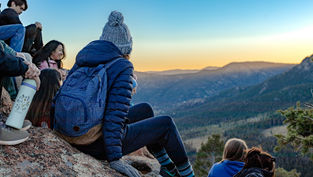 People sitting on a mountain ledge at sunset, wearing winter clothing. Blue sky, distant mountains, and a relaxed, contemplative mood.