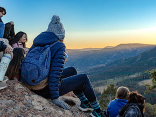 People sitting on a mountain ledge at sunset, wearing winter clothing. Blue sky, distant mountains, and a relaxed, contemplative mood.