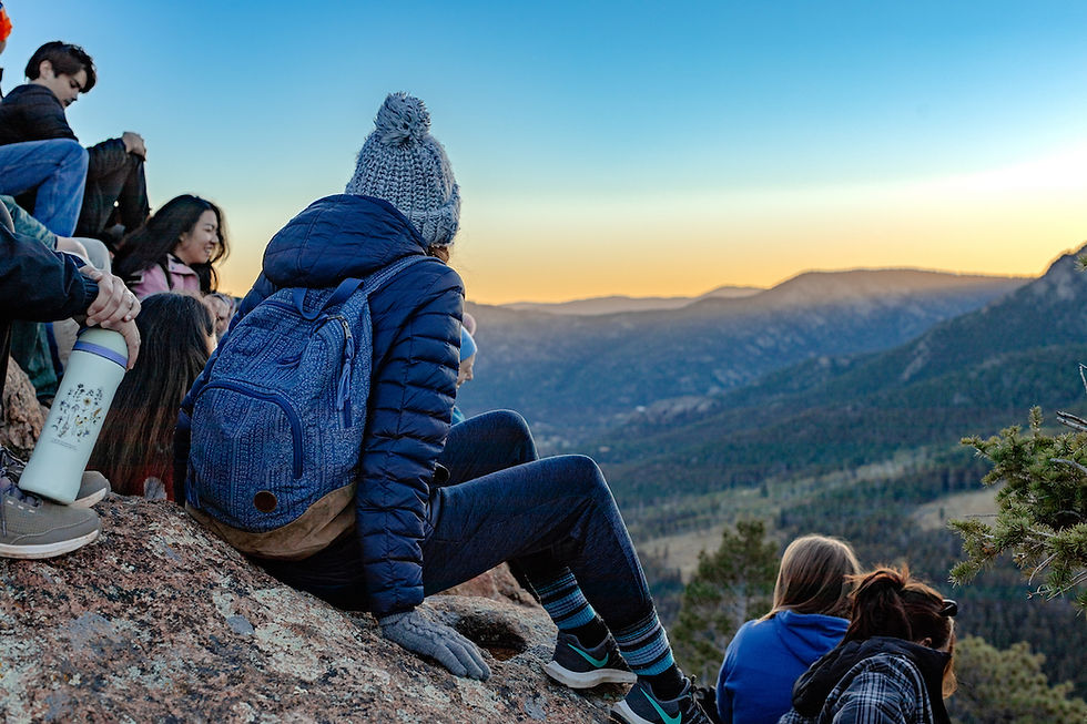 People sitting on a mountain ledge at sunset, wearing winter clothing. Blue sky, distant mountains, and a relaxed, contemplative mood.