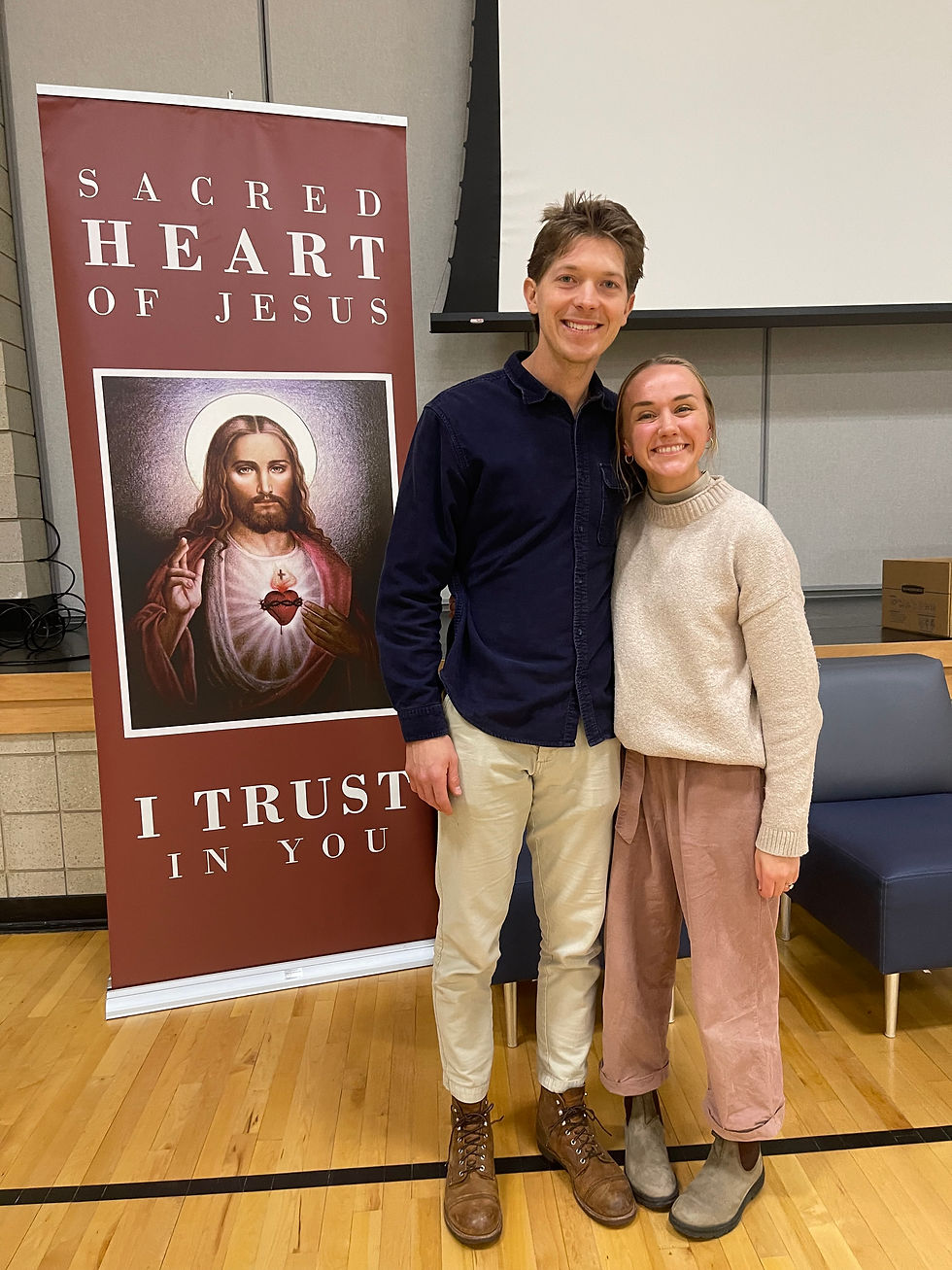 Two people smiling in front of a maroon banner with "Sacred Heart of Jesus, I Trust in You" text. Indoor setting, wooden floor.