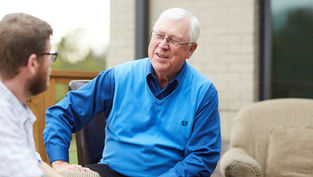 Elderly man in blue vest smiling while talking to another person outdoors, with a brick wall and porch furniture in the background.