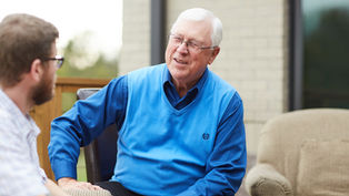 Elderly man in blue vest smiling while talking to another person outdoors, with a brick wall and porch furniture in the background.
