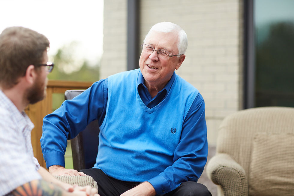 Elderly man in blue vest smiling while talking to another person outdoors, with a brick wall and porch furniture in the background.