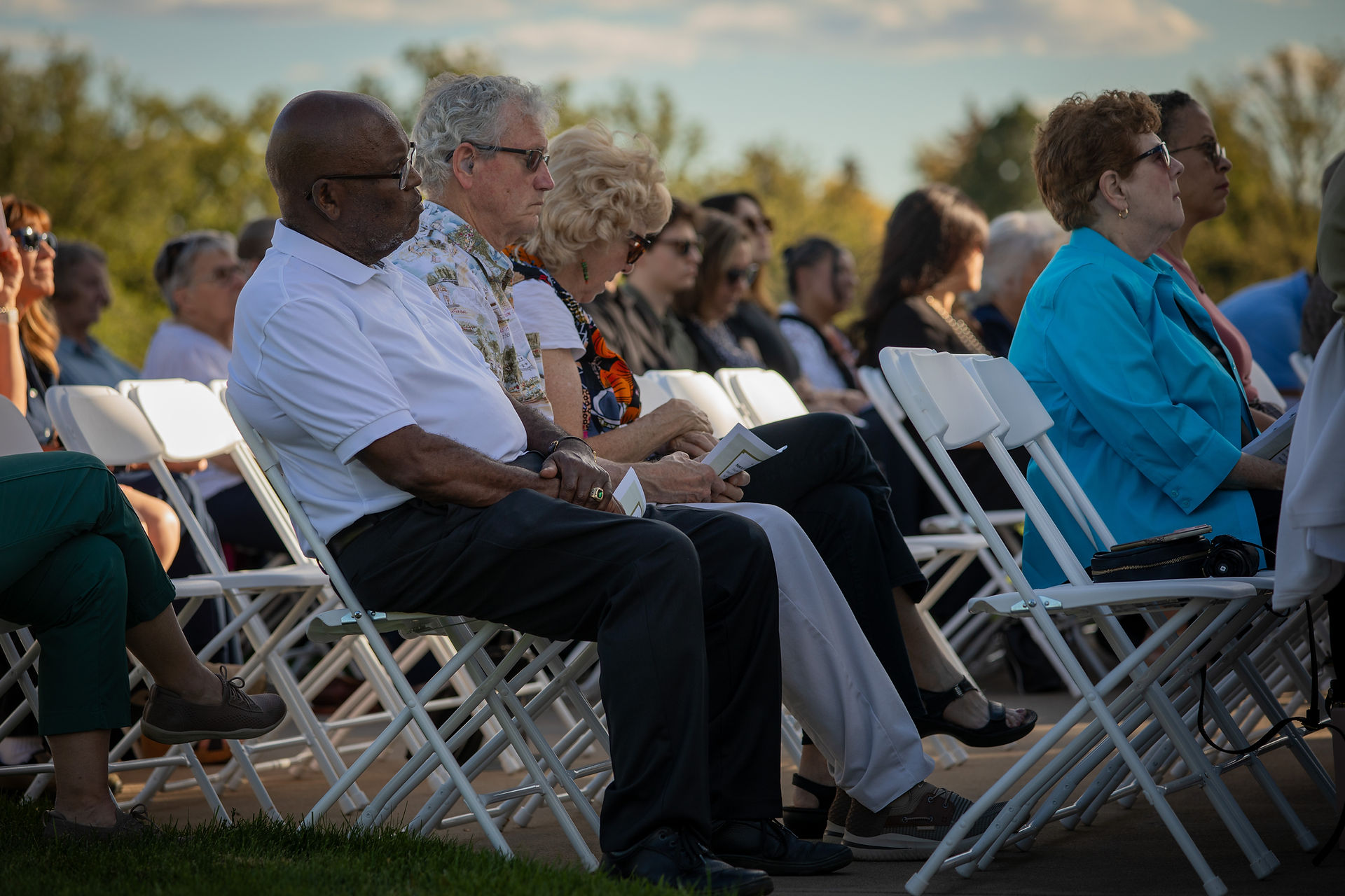 Audience seated outdoors in rows of white chairs, attentively watching an event. Trees and a partly cloudy sky in the background.