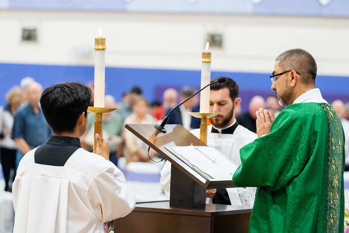 Deacon in green vestments prays at church lectern while two acolytes hold lit candles, with congregation blurred behind.