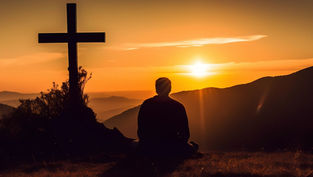 Silhouette of person sitting near a cross, gazing at a vibrant orange sunset over hills, creating a serene and contemplative mood.