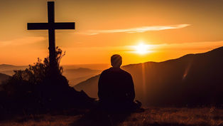 Silhouette of person sitting near a cross, gazing at a vibrant orange sunset over hills, creating a serene and contemplative mood.